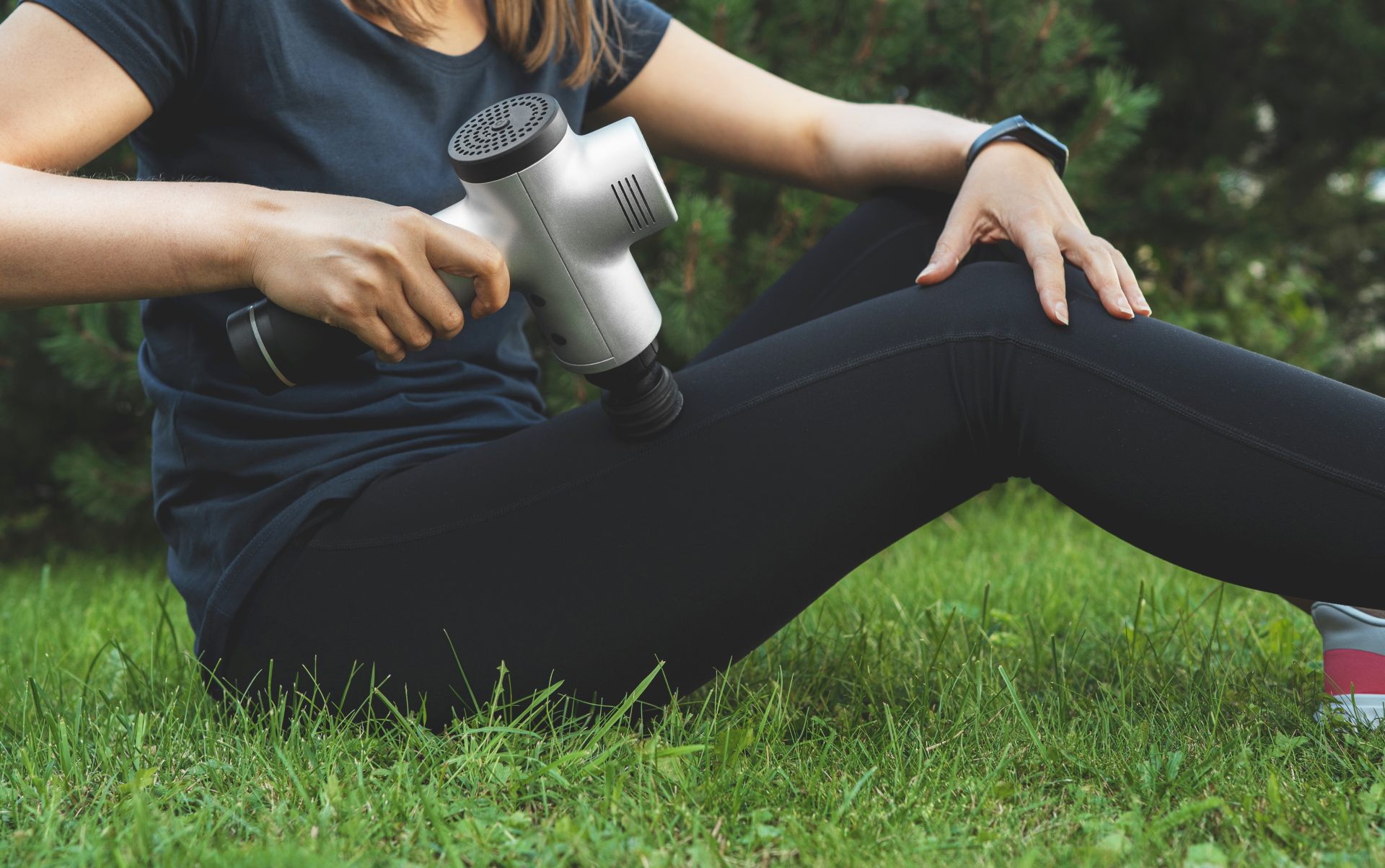 A woman in black workout clothing uses a percussion massager on her leg while sitting on the grass