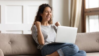 A smiling woman cheers while sitting on her soft with her laptop perched on her legs, representing class action settlements closing soon.