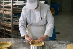 A pastry chef kneeding pie crust with pie racks sitting in the background. The stainless steel table is covered in flour dust.