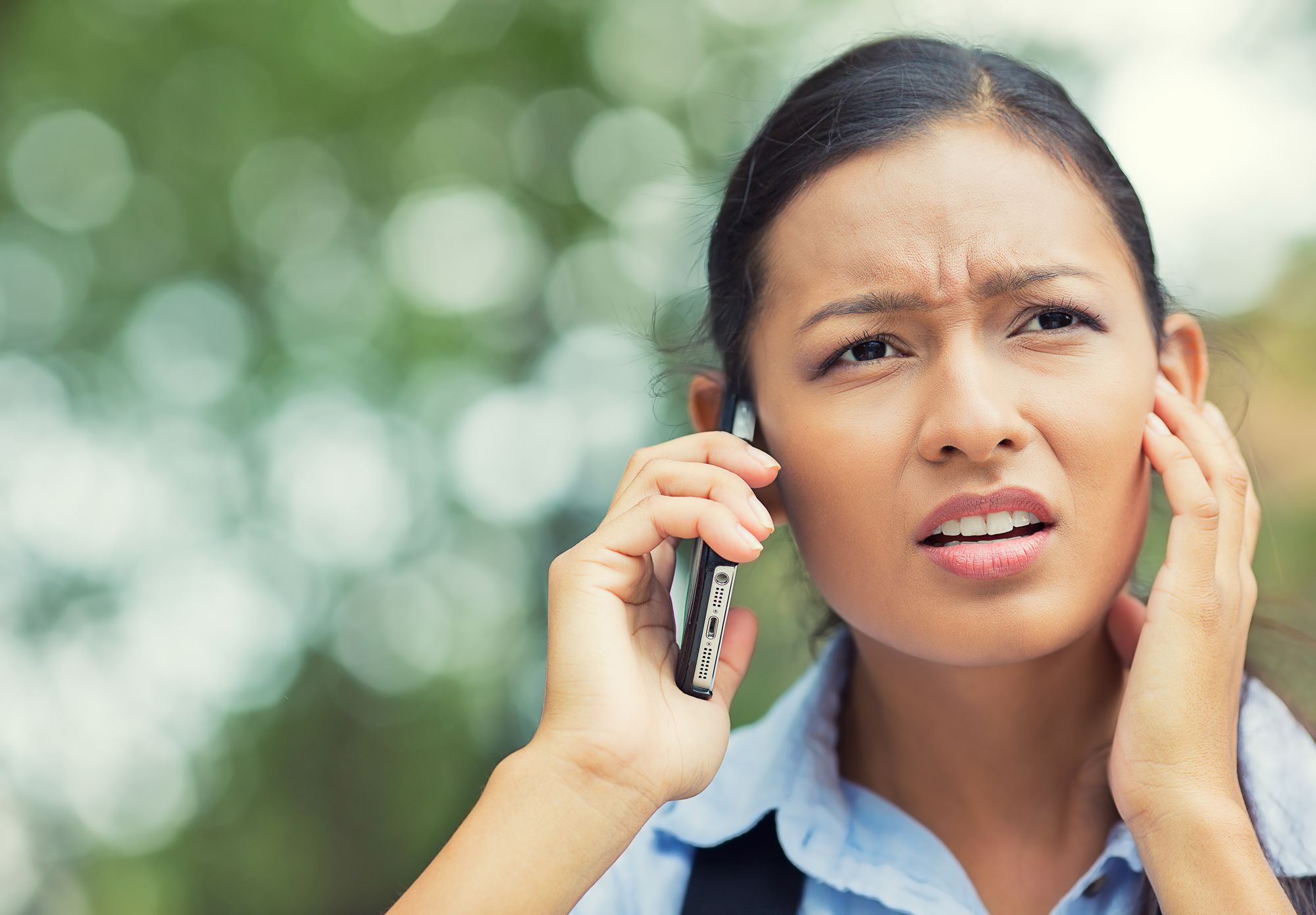 Closeup portrait, headshot Frustrated young businesswoman in formalwear talking on mobile phone, touching face, closing ear with hand while standing outdoors, bad news, cellular connection concept