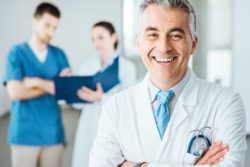 Confident doctor posing and smiling at camera and medical staff checking medical records on background