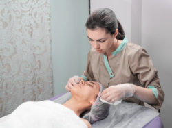 Spa therapy for young woman receiving facial mask at beauty salon. Beautician and client before Facial Mask in beauty salon. Beautician worker applying facial cosmetic mask to client in beauty salon.