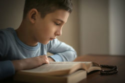 A boy sits at a table with a bible and a rosary.