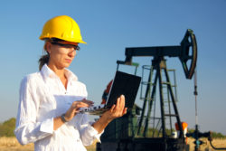 A woman works a laptop next to an oil drill.