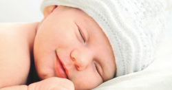 A smiling baby sleeps wearing a cap.