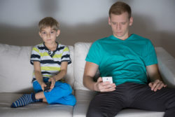 A boy watches TV while his dad looks at a phone.