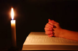A candle illuminates hands clasped over a bible.