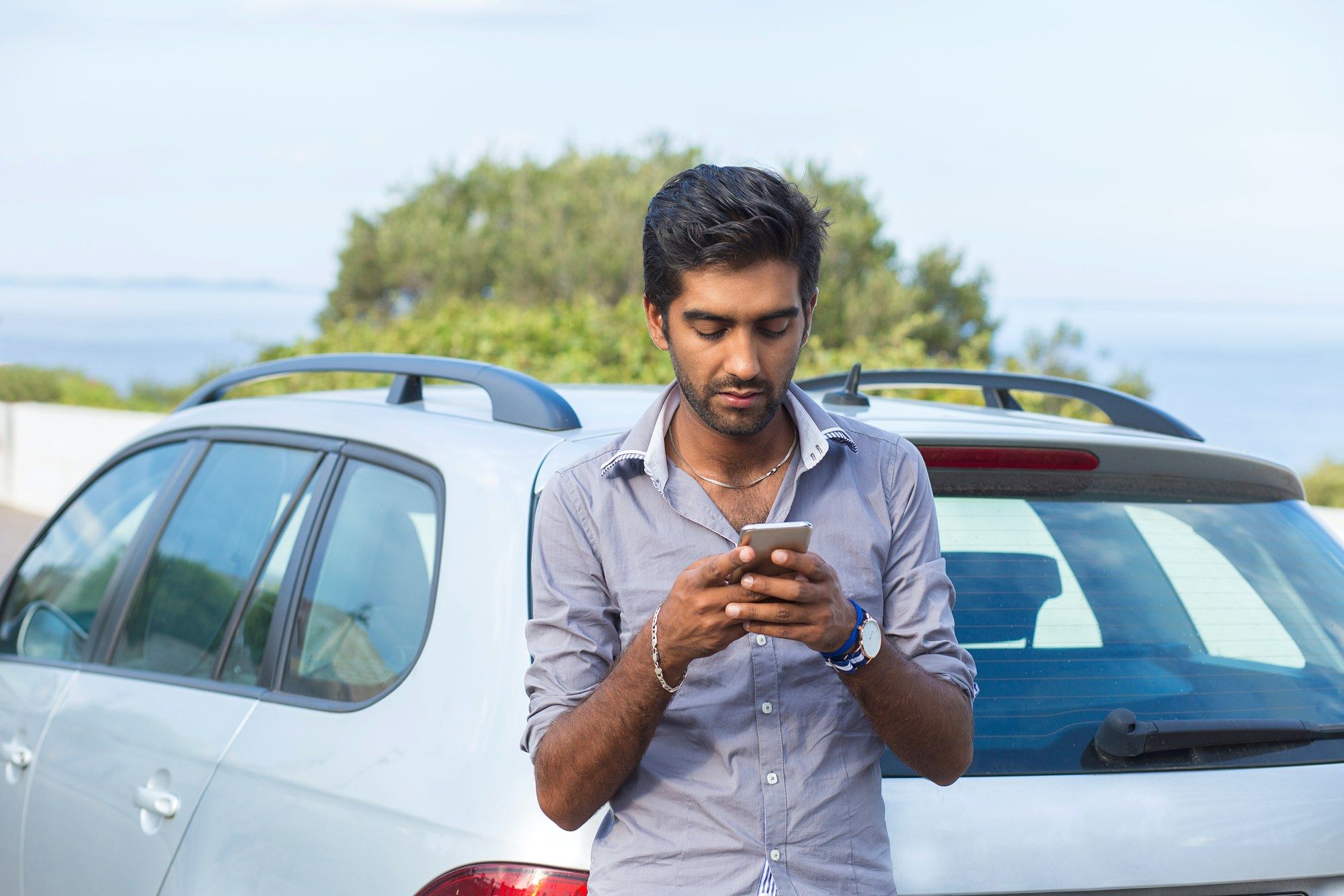 Young-man-leans-on-car-reading-text-on-smartphone