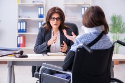 A woman in a wheelchair, neck brace, and sling talks with a woman sitting behind a desk - Long term disability claims
