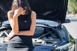 Woman holds her head in her hand as she leans against broken down car on side of road