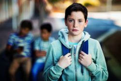 Sad schoolboy stands wearing sweatshirt and backpack.