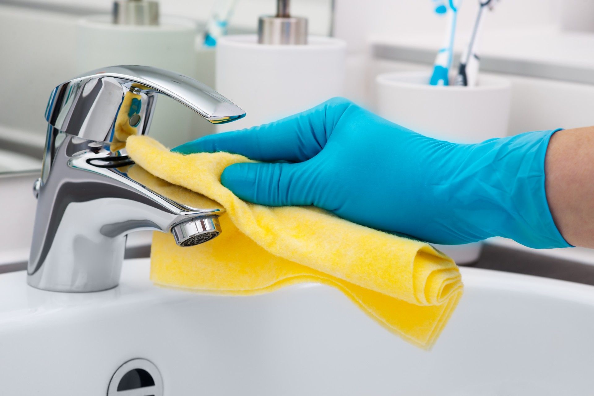 Person wearing a blue rubber glove, cleaning a bathroom sink faucet with a yellow cloth