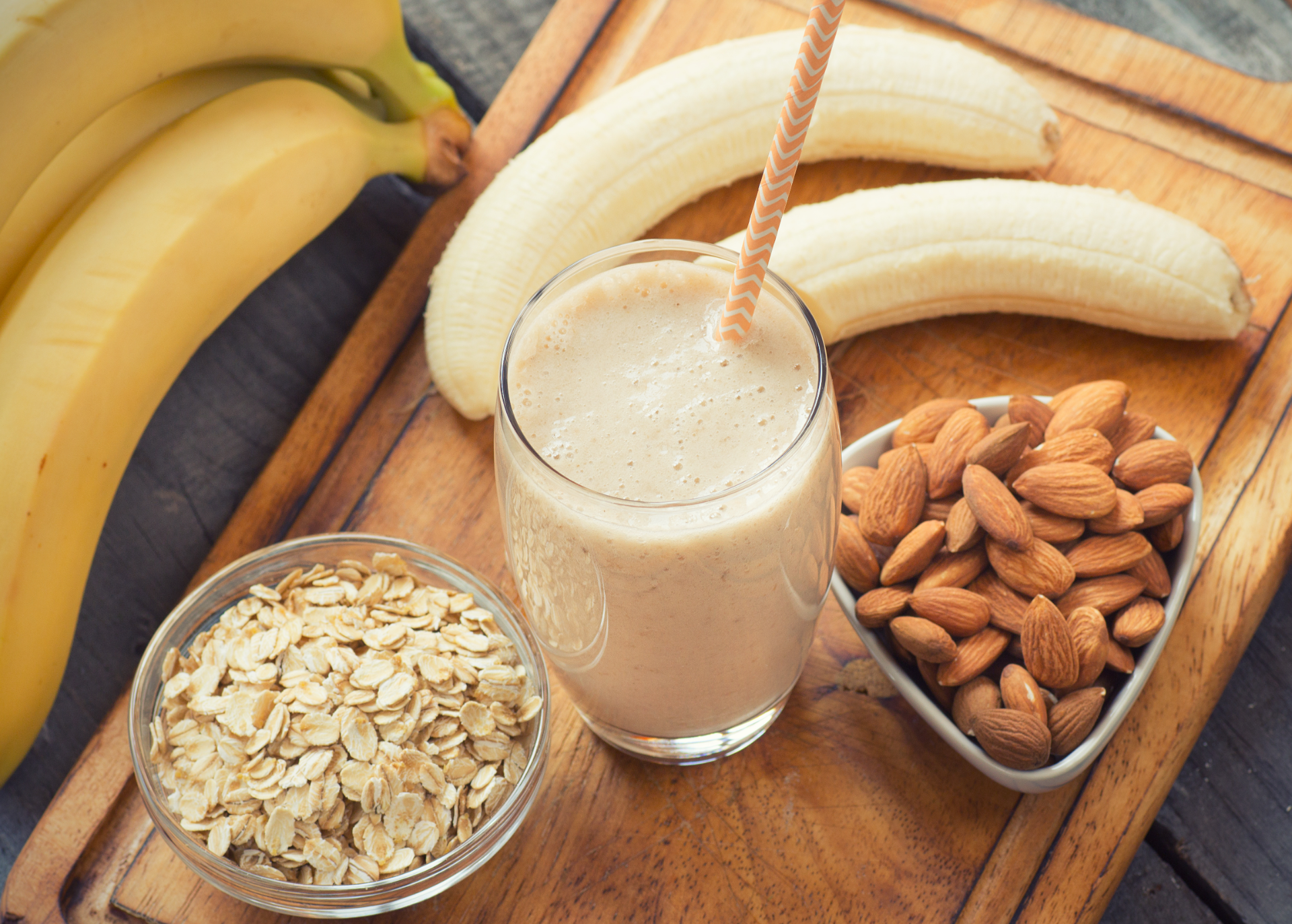 Smoothie in a glass with straw, sitting on a cutting board and surrounded by bananas, oats and almonds