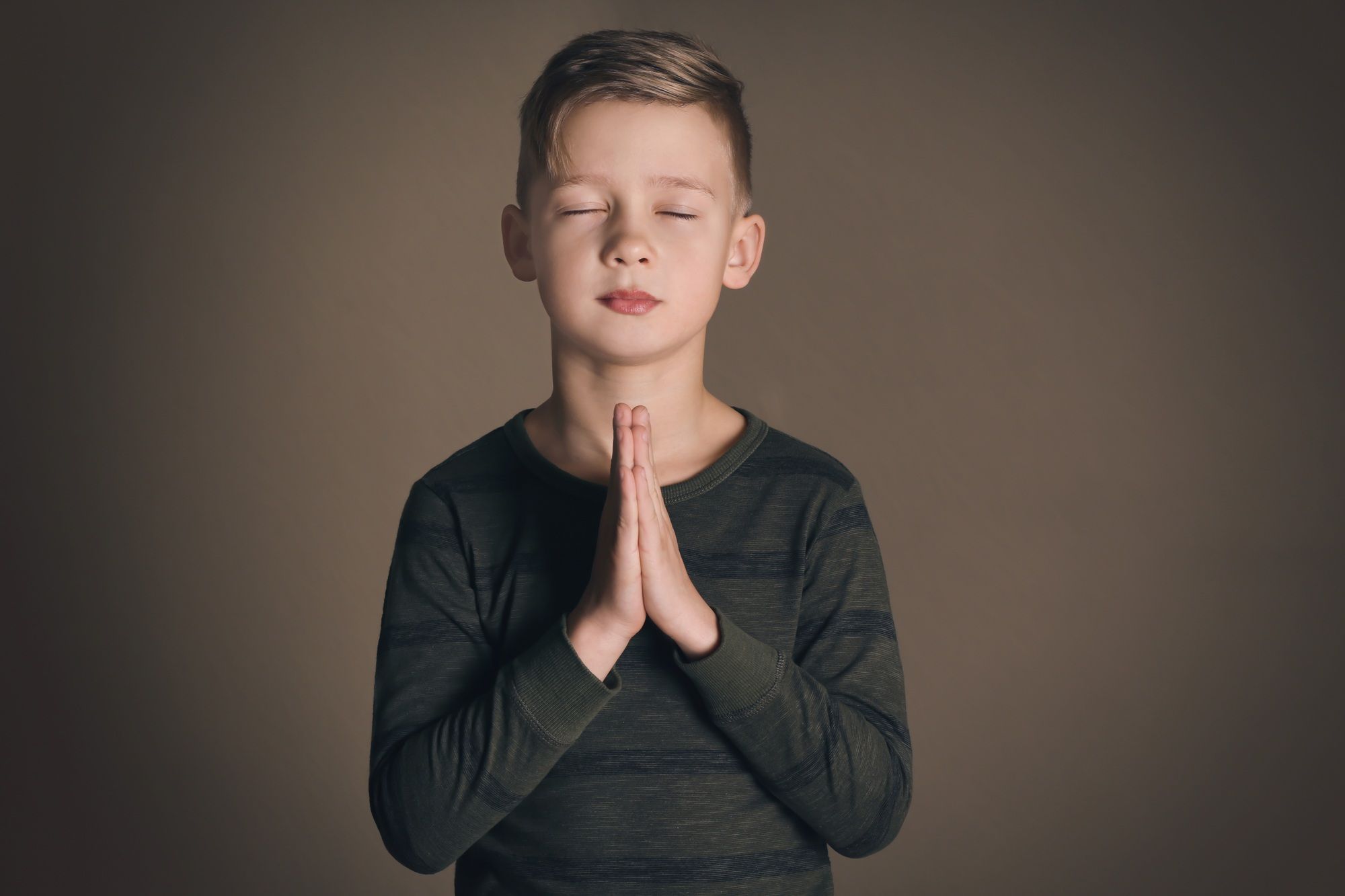 Young boy faces camera with eyes closed and hands folded in prayer