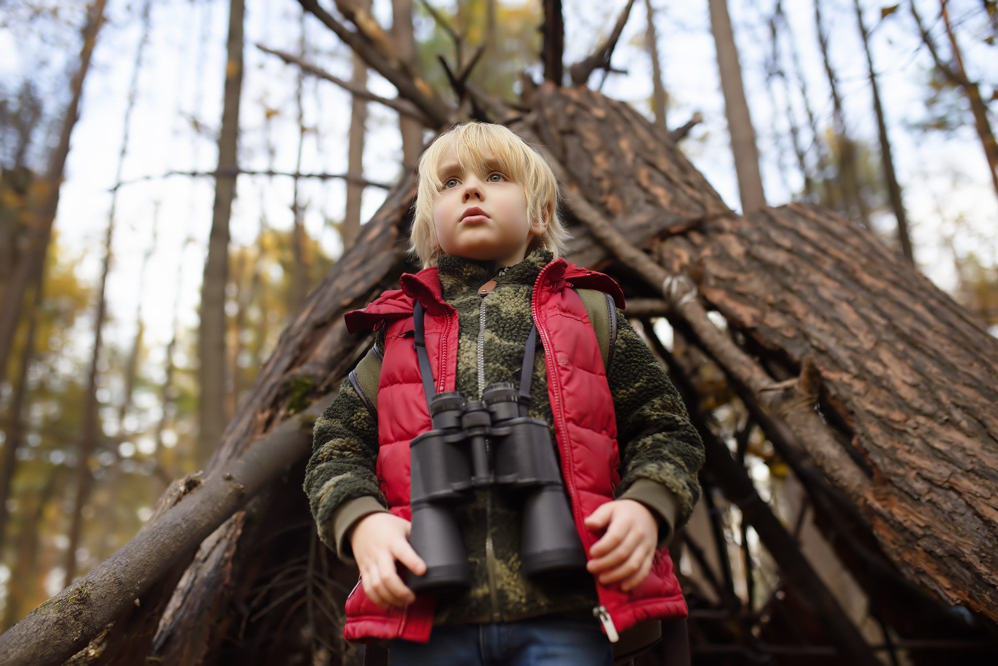 Young boy with binoculars stands in front of teepee tent in the woods