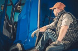 Male truck driver sits next to his blue semi truck 