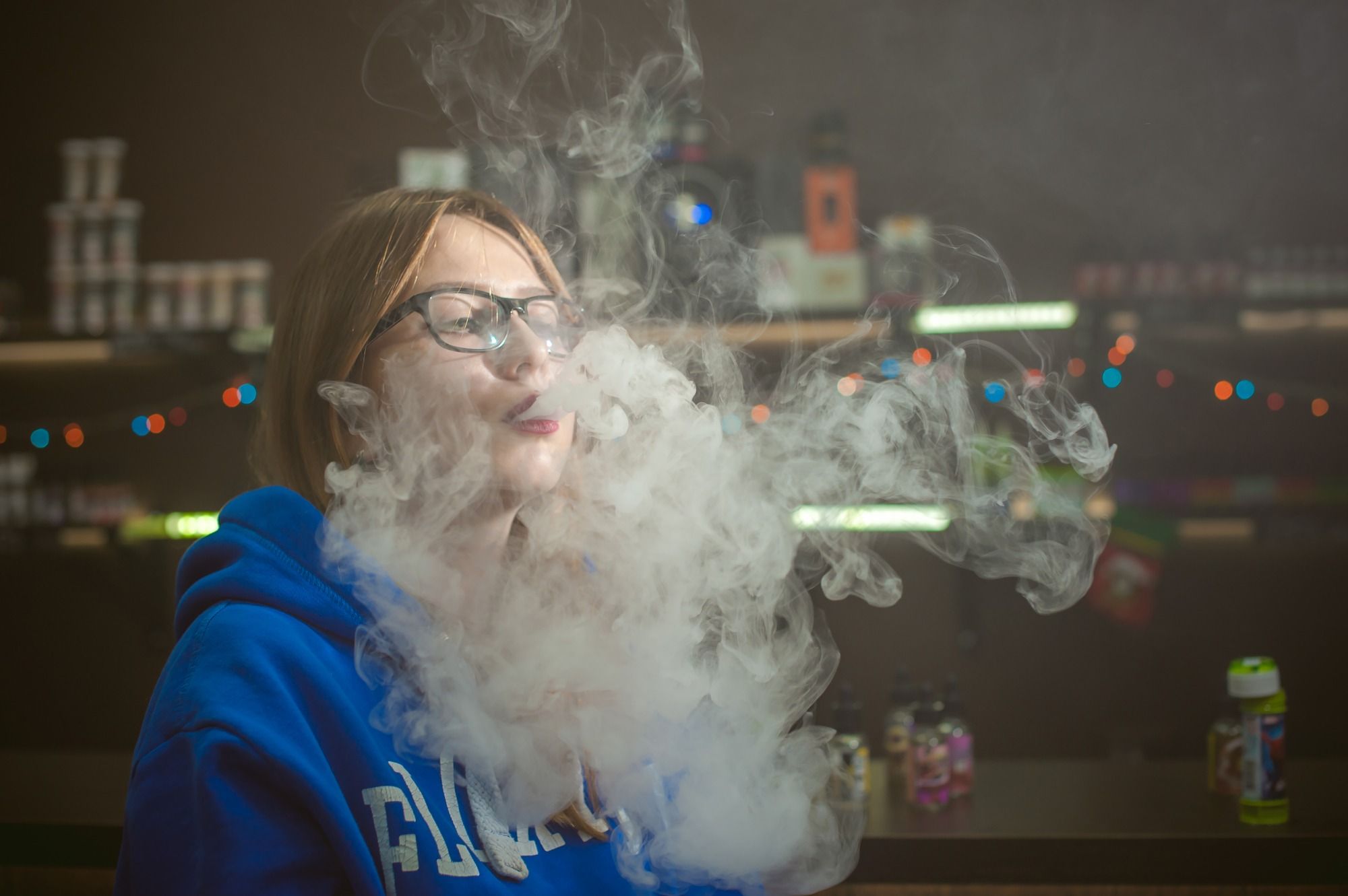 Woman in glasses and blue sweatshirt exhales vaping cloud