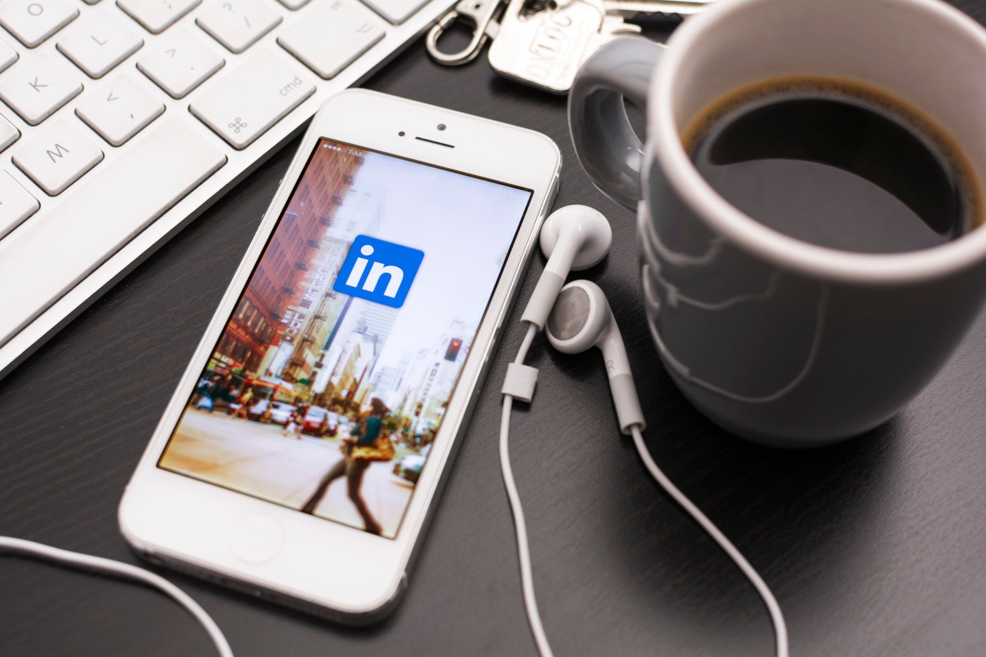 An iPhone displays the LinkedIn app while lying next to coffee mug, keyboard and earbuds