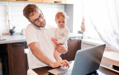 Man in kitchen holds baby, talks on phone and works on laptop - paid leave