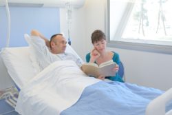 ill man in hospital bed with woman reading to him
