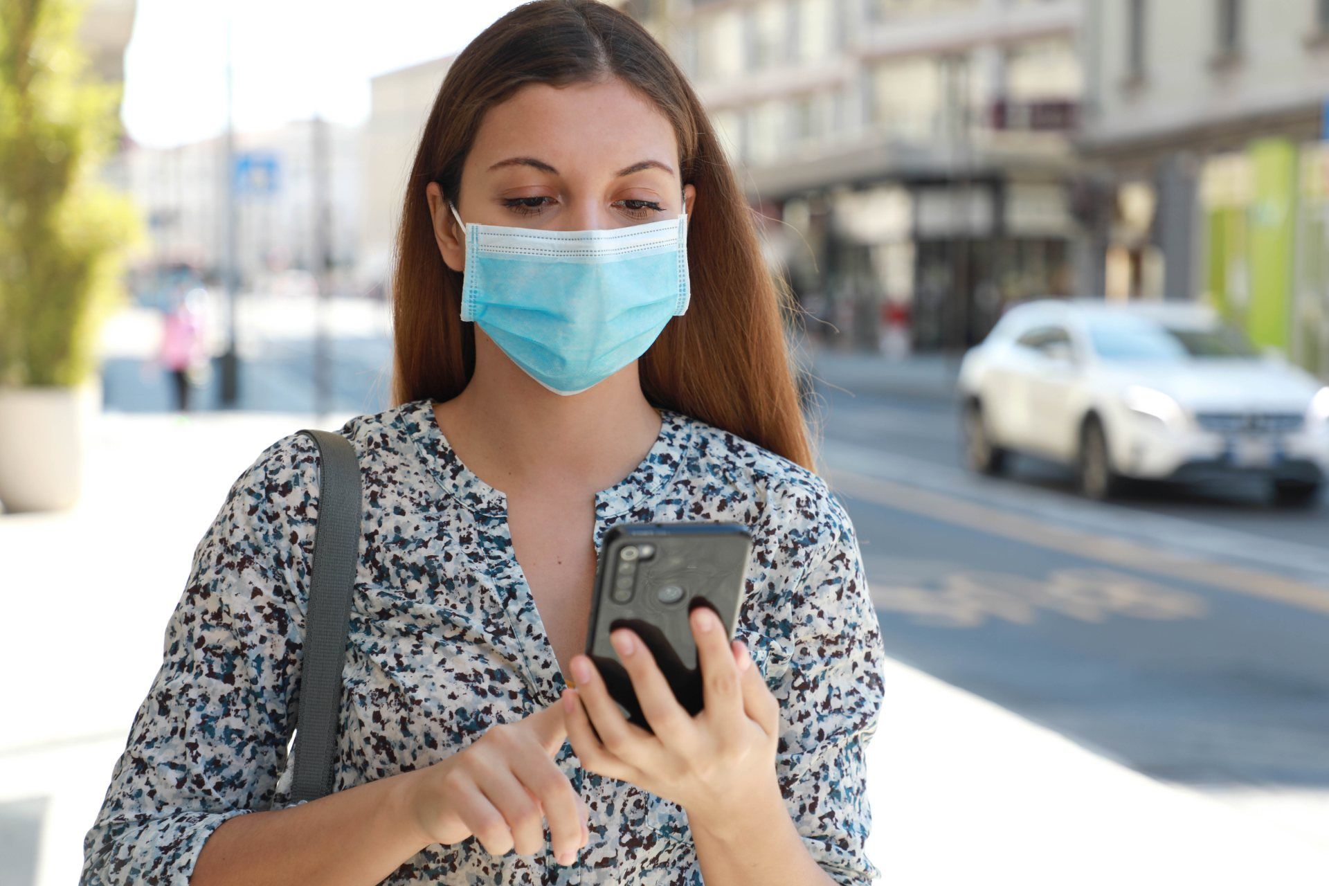 Woman in surgical mask stands on city street, using a smartphone - coronavirus tracker app