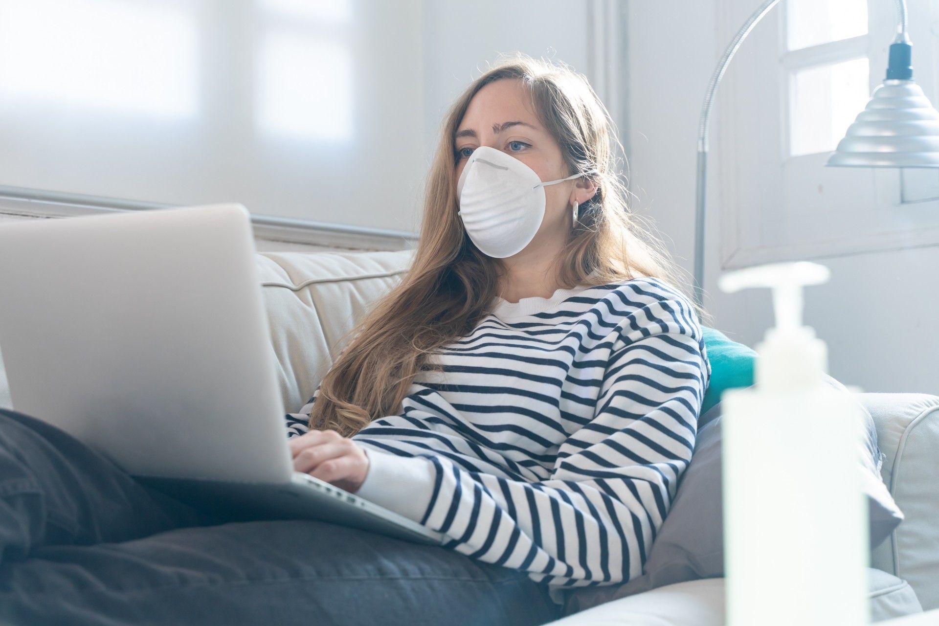 A woman lies on her couch, working from home on a laptop while wearing a face mask near a bottle of hand sanitizer - paid leave