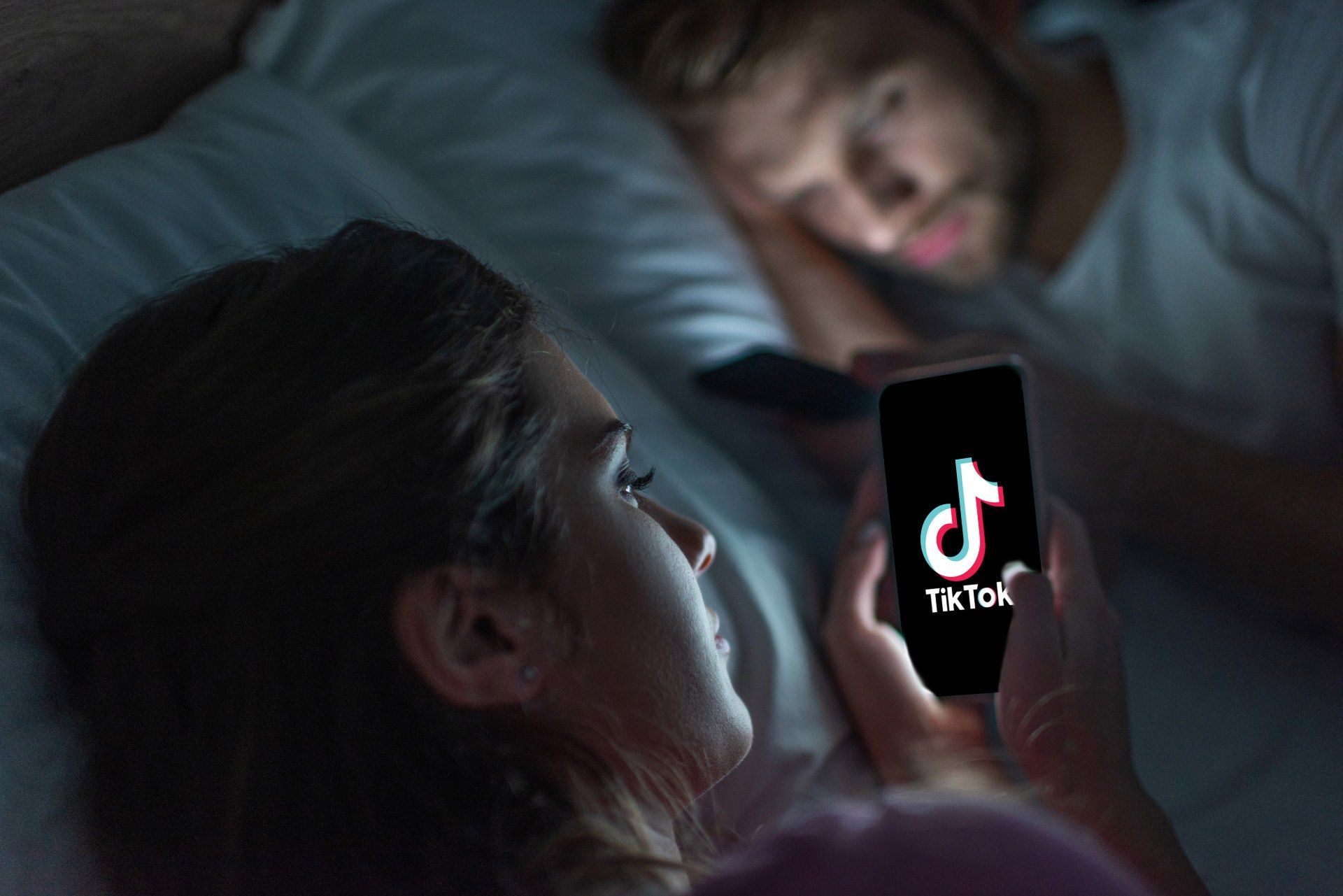 Woman lies in bed looking at her smartphone, which shows the TikTok app load screen