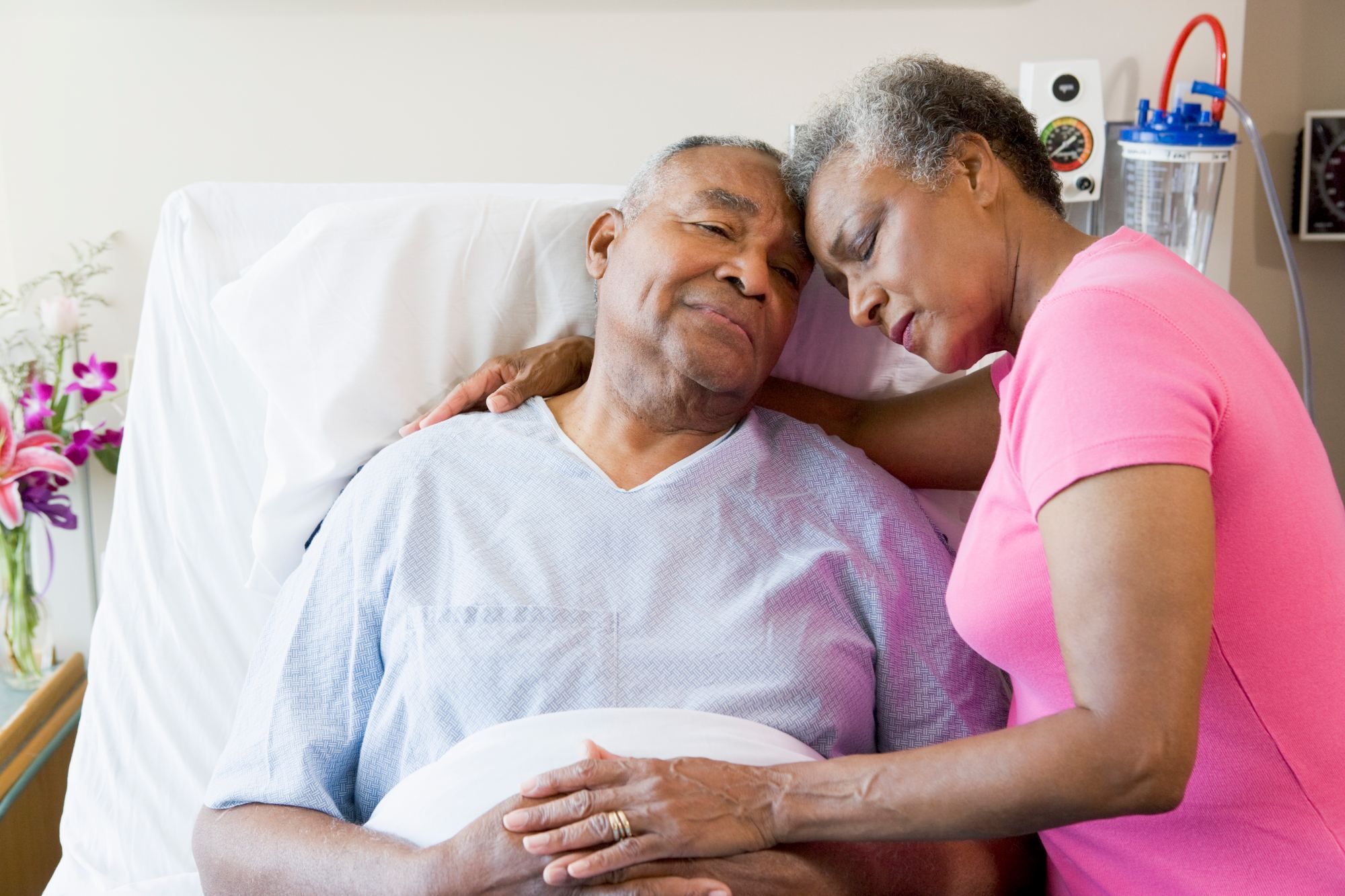 Elderly couple hug while man lies in hospital bed