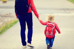 Mom holds hands with daughter with backpack as they walk along sidewalk
