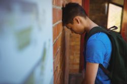 Sad schoolboy wearing backpack leans his head against a brick wall