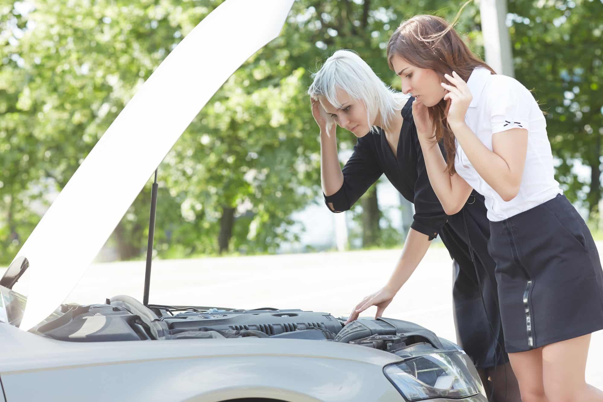 Two women look under hood of broken down sedan