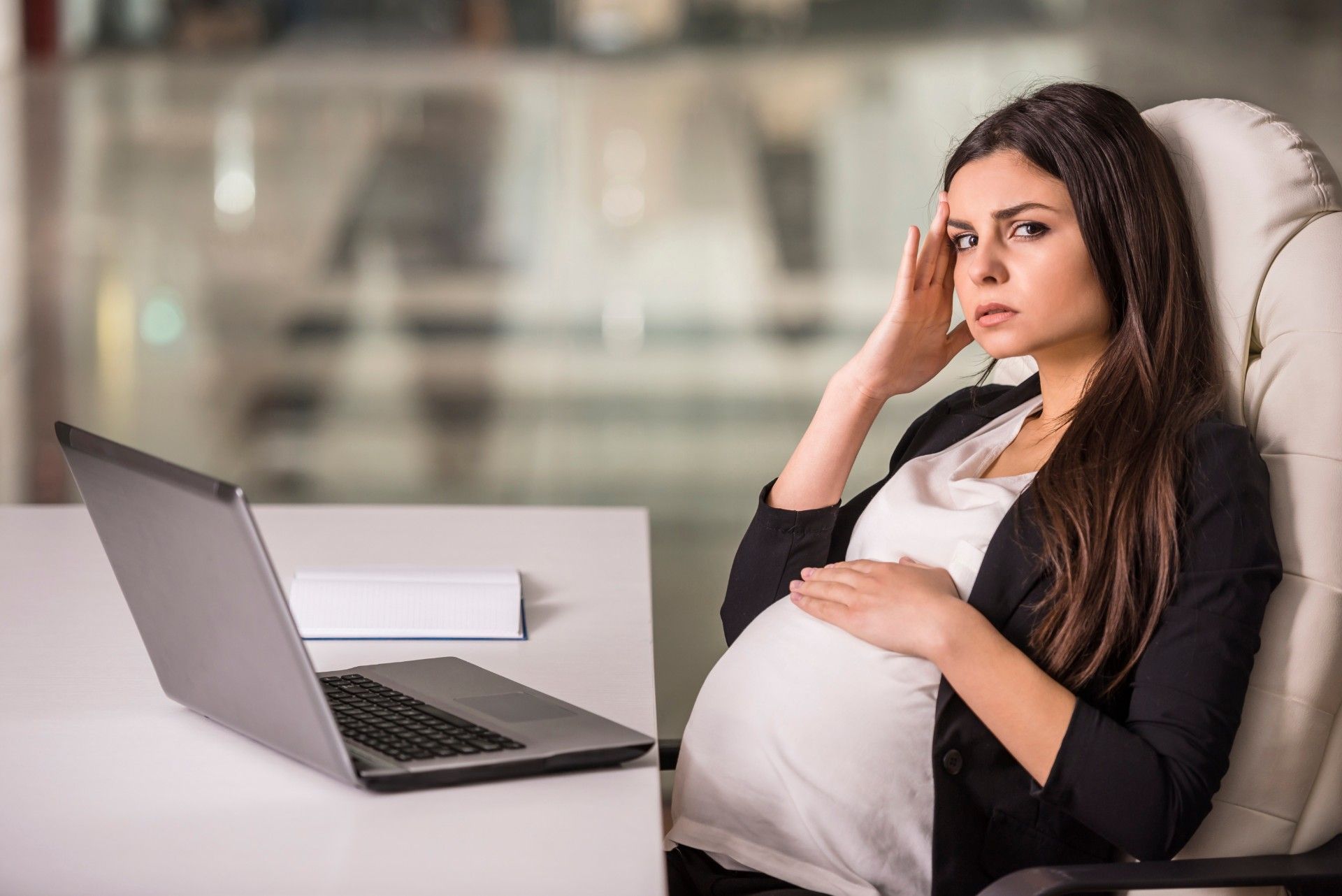 An annoyed pregnant woman sits at a table with a laptop in an office - pregnant worker rights