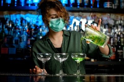A female bartender in a mask pours a green drink into one of three coupe glasses - pandemic rules