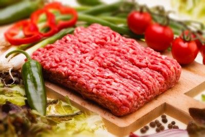 A rectangle of ground beef sits on a wood cutting board surrounded by vegetables - beef prices