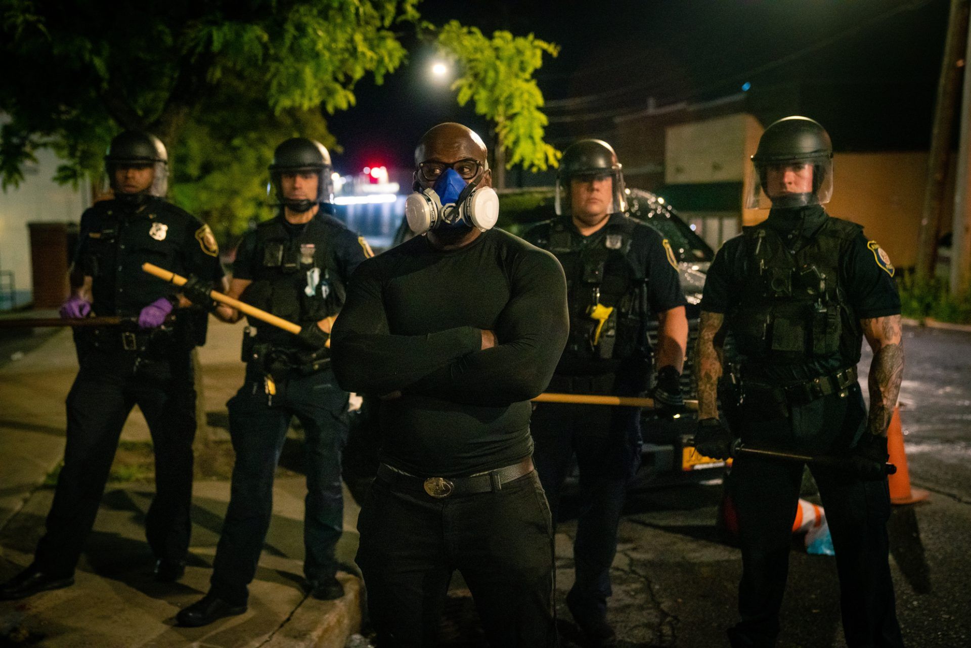 A Black man wearing a gas mask stands in front of a line of police wearing riot gear, facing away - police brutality