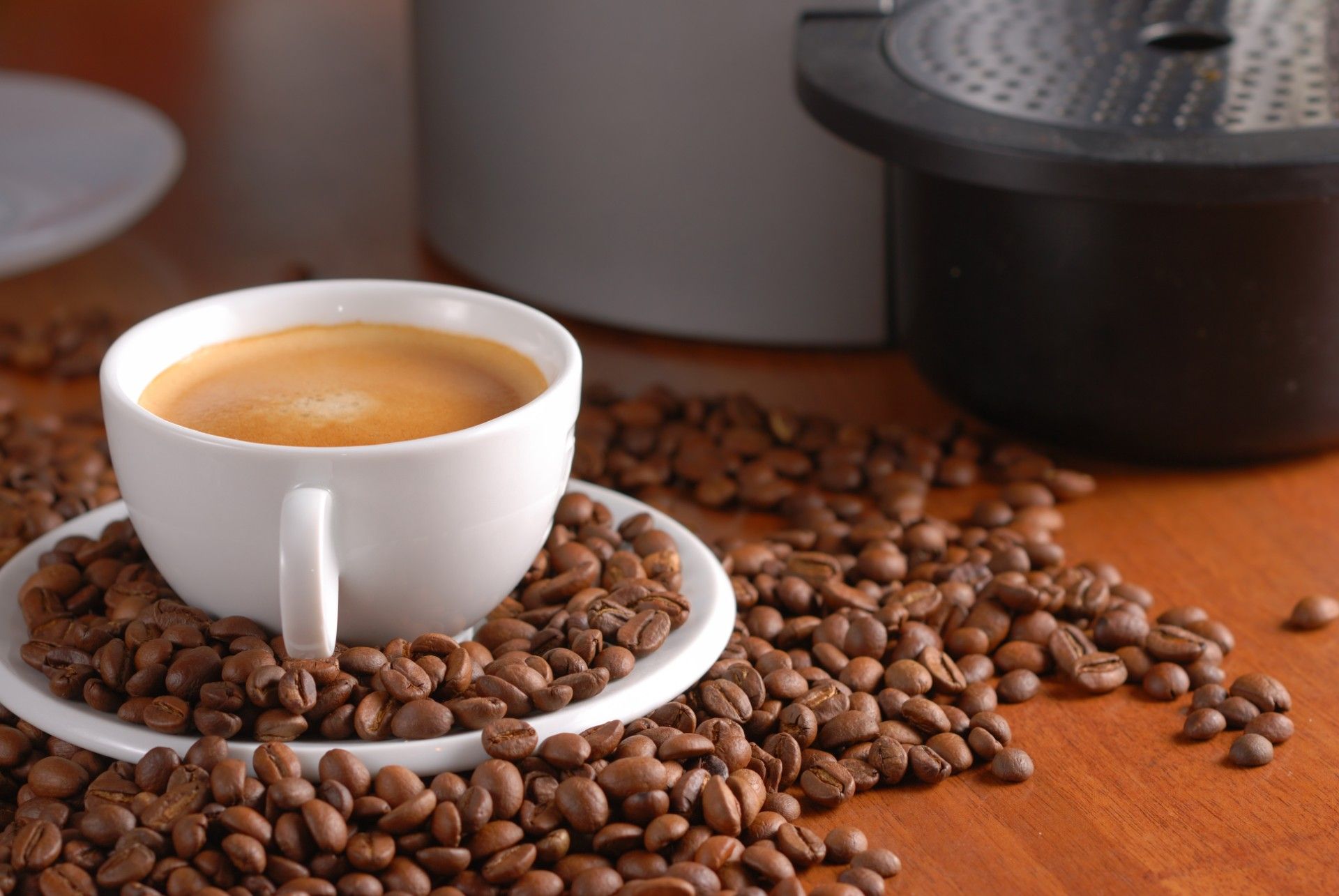 A cup of coffee surrounded by coffee beans sits near a coffee maker - keurig coffee pods
