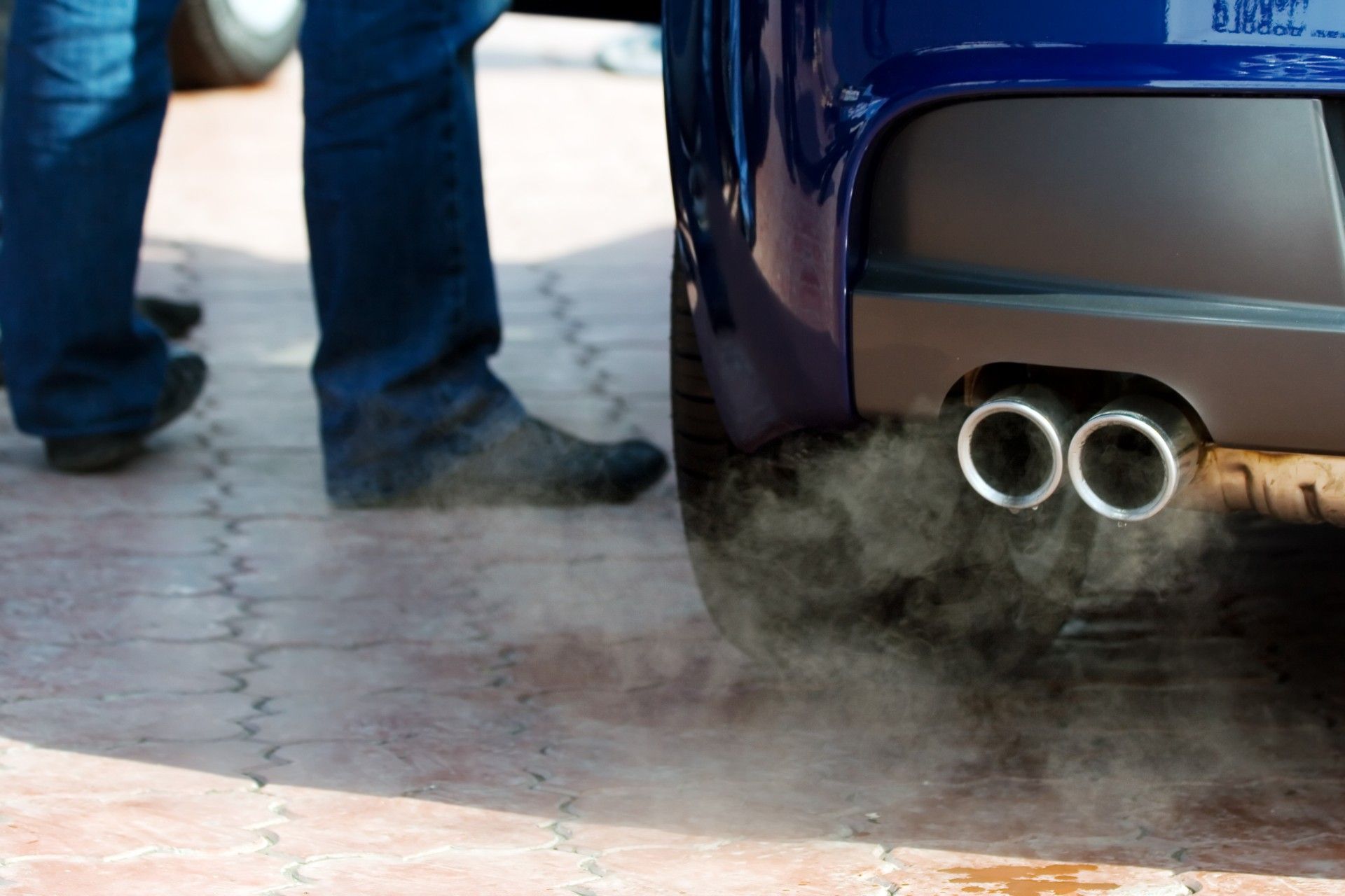 Low shot of a person's legs near a vehicle that has exhaust coming from the exhaust pipe - vehicle emissions