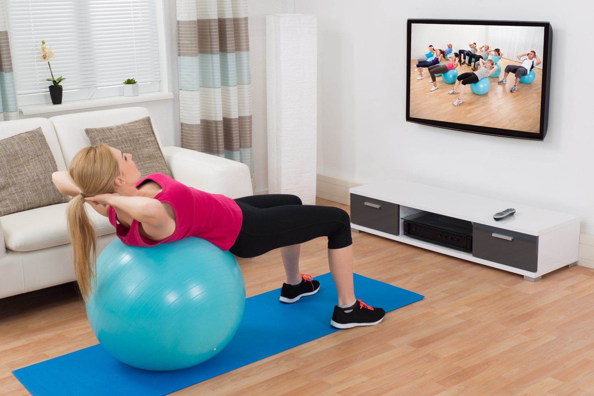 A woman uses an exercise ball on a blue yoga mat while following along with a workout video - Beachbody workout videos