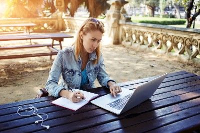A female student studies at a picnic table with a laptop, notebook and earbuds - college tuition
