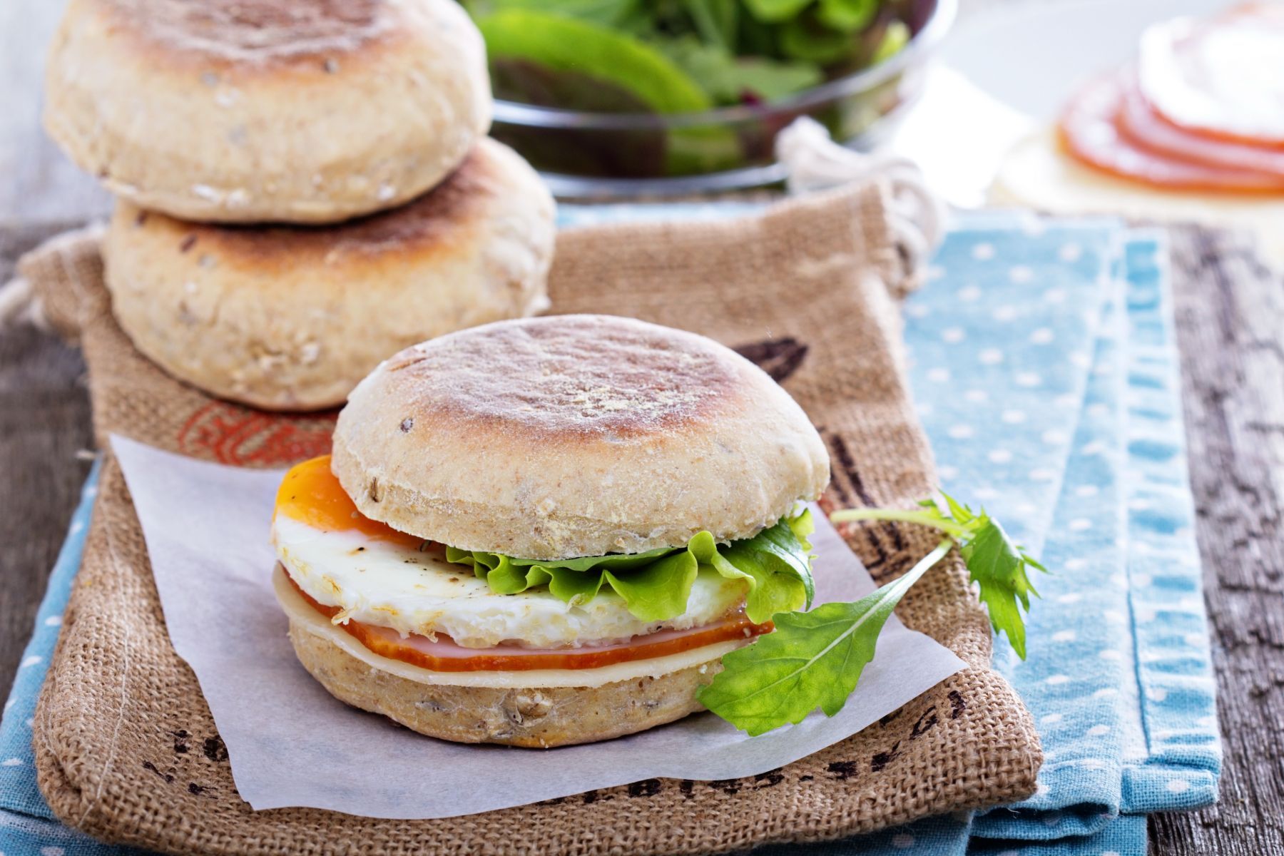 English muffin breakfast sandwich sits near two other English muffins on a table with cloth napkins - whole-grain foods