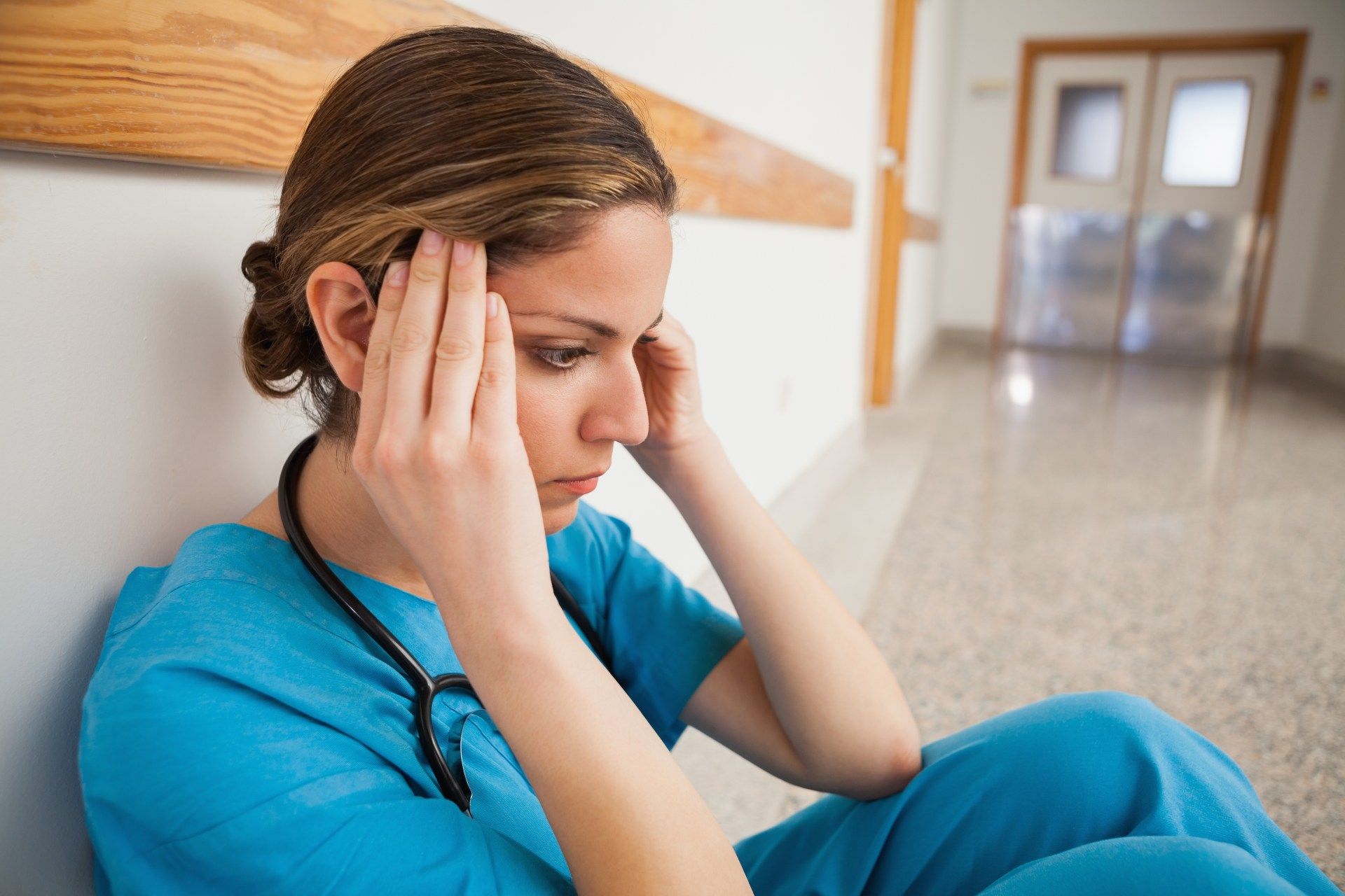 A nurse in blue scrubs sits on a hospital hallway floor, rubbing her temples - coronavirus outbreak