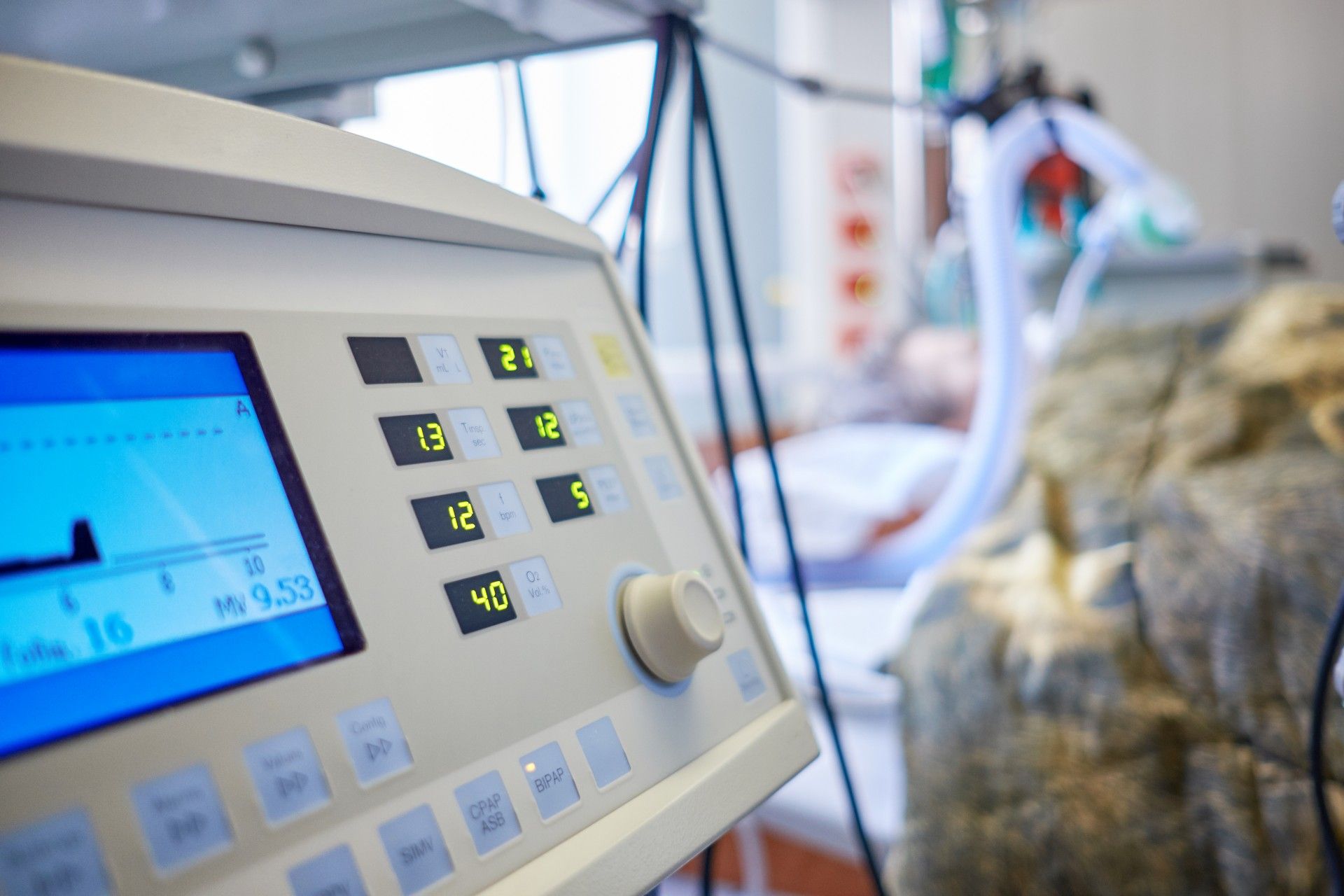 A ventilator in the foreground with a patient hooked up to it lying in a bed in the background - ventilators