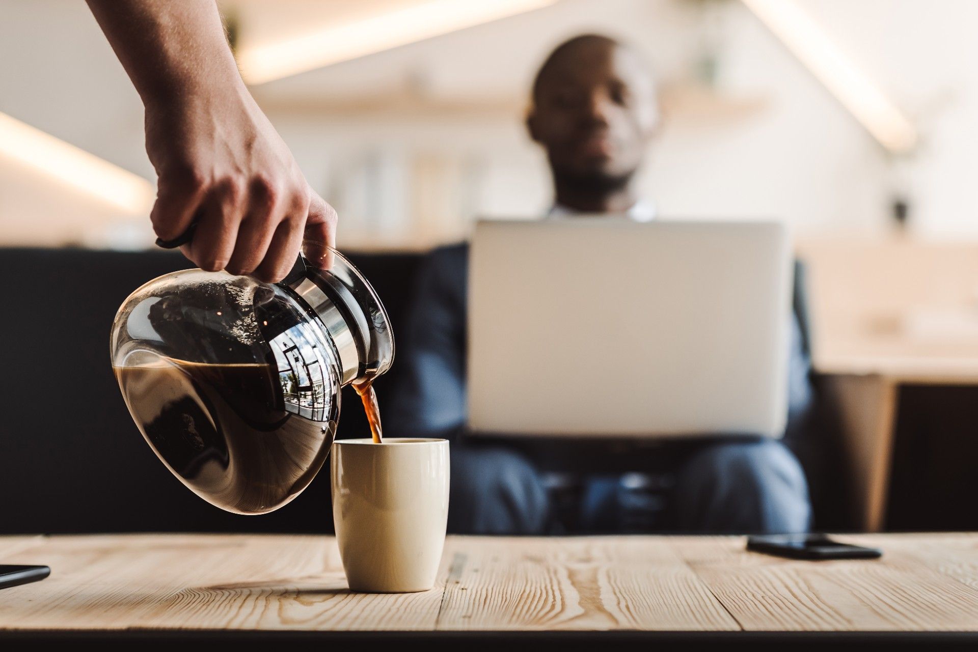 Someone pours coffee for man sitting near a table while working on a laptop - Maxwell House