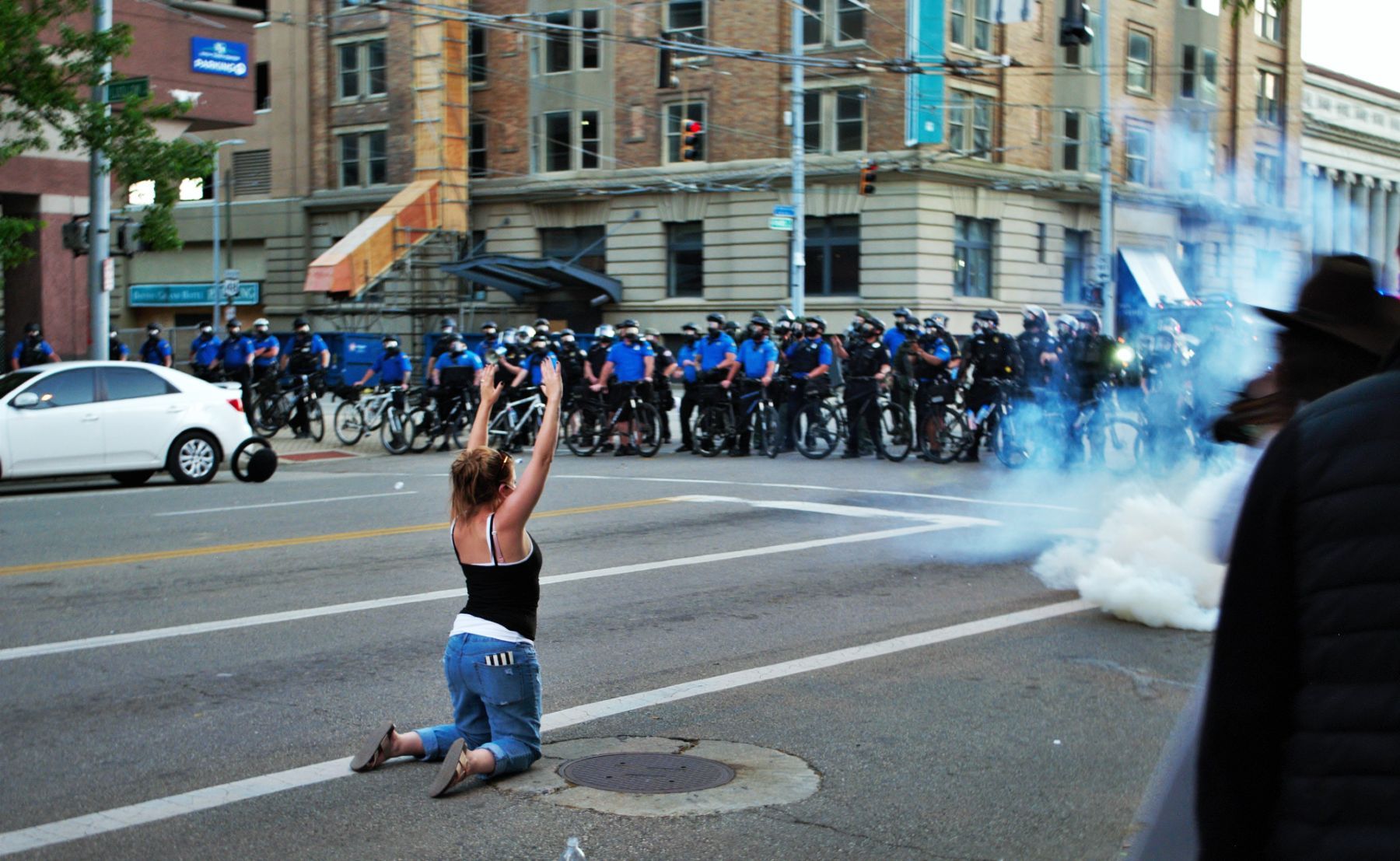 Police officers deploy tear gas at a protest as a protester kneels in the street with her hands raised - racial discrimination