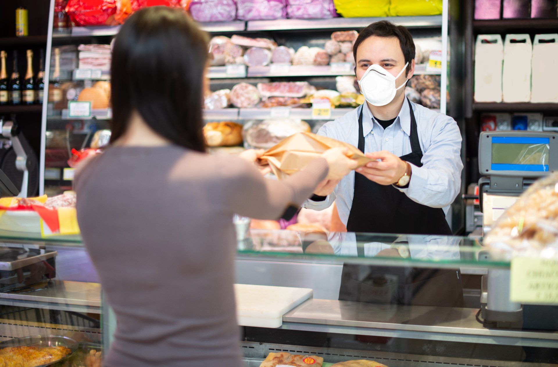 A deli counter worker wearing a surgical mask hands a woman her purchase - employee safety