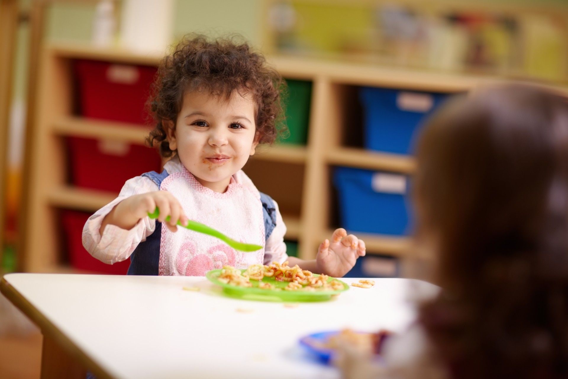 A happy young child in a bib eats while sitting at a classroom table - Feeding Our Future