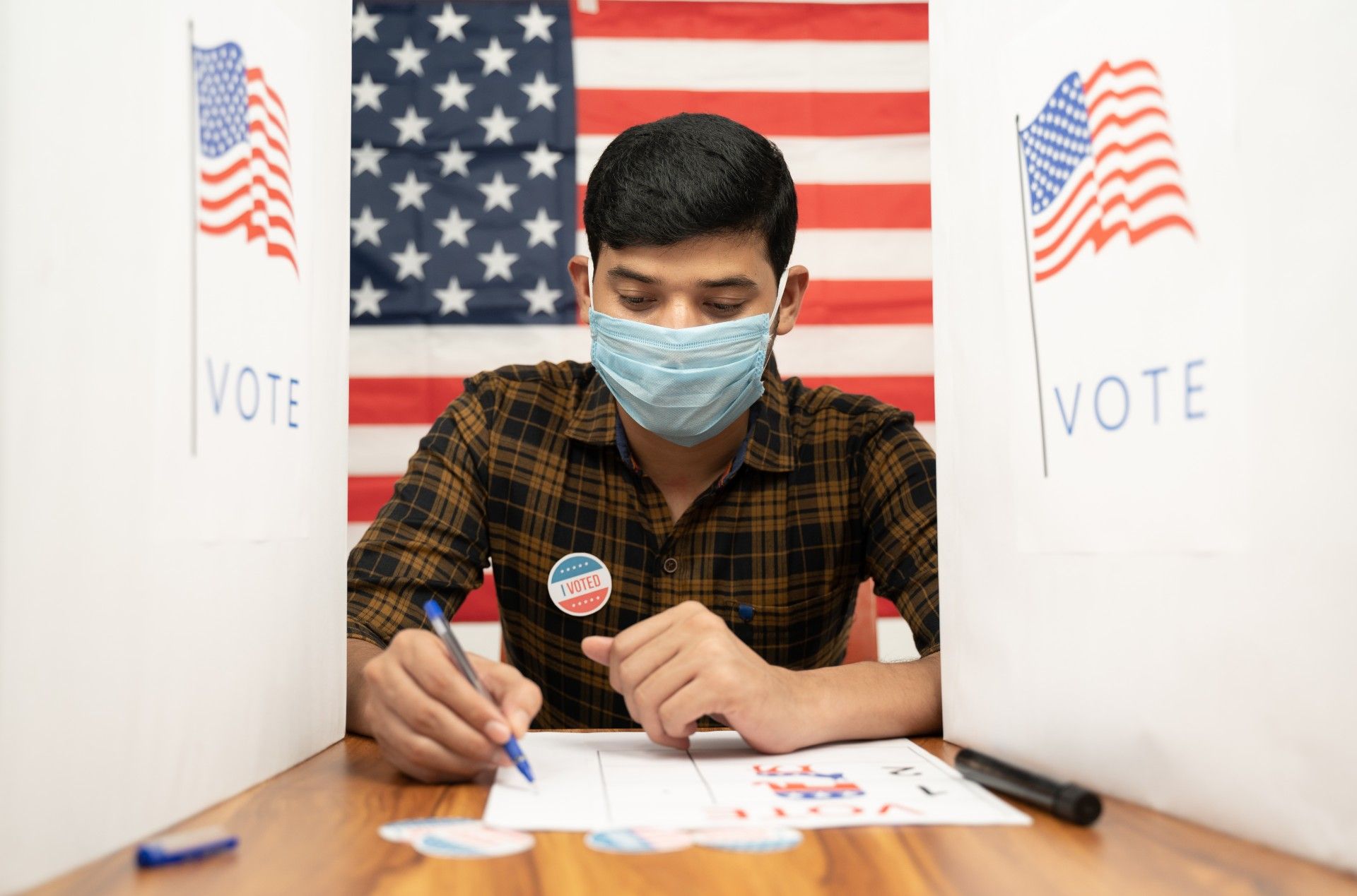 A man sits in a polling booth to vote while wearing a mask, with an American flag backdrop - arkansas voters