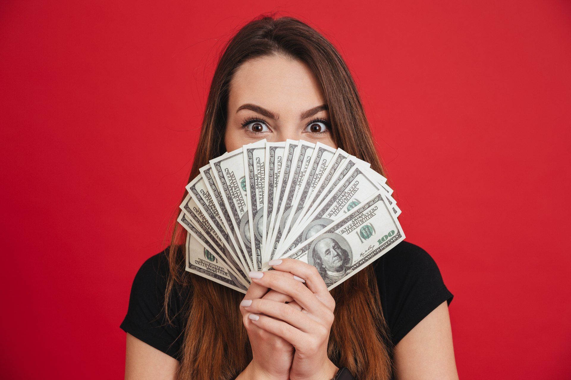 A woman in a black shirt holds fanned-out cash in front of the lower half of her face against a red background