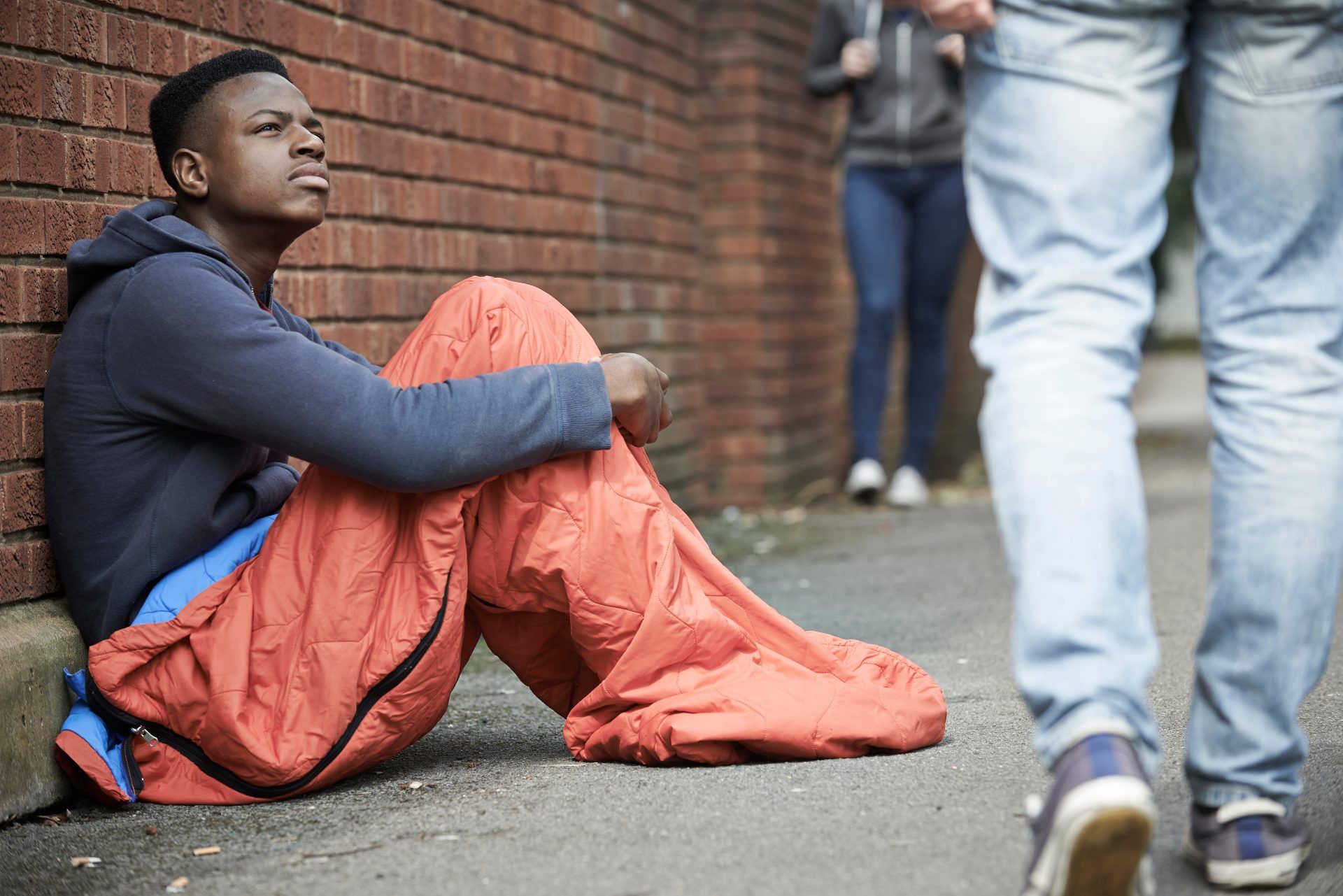 A homeless young man sits on a sidewalk with his legs inside an orange sleeping bag - homeless youth