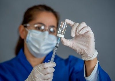 A woman in a surgical mask, goggles, blue scrubs and rubber gloves fills a syringe with vaccine